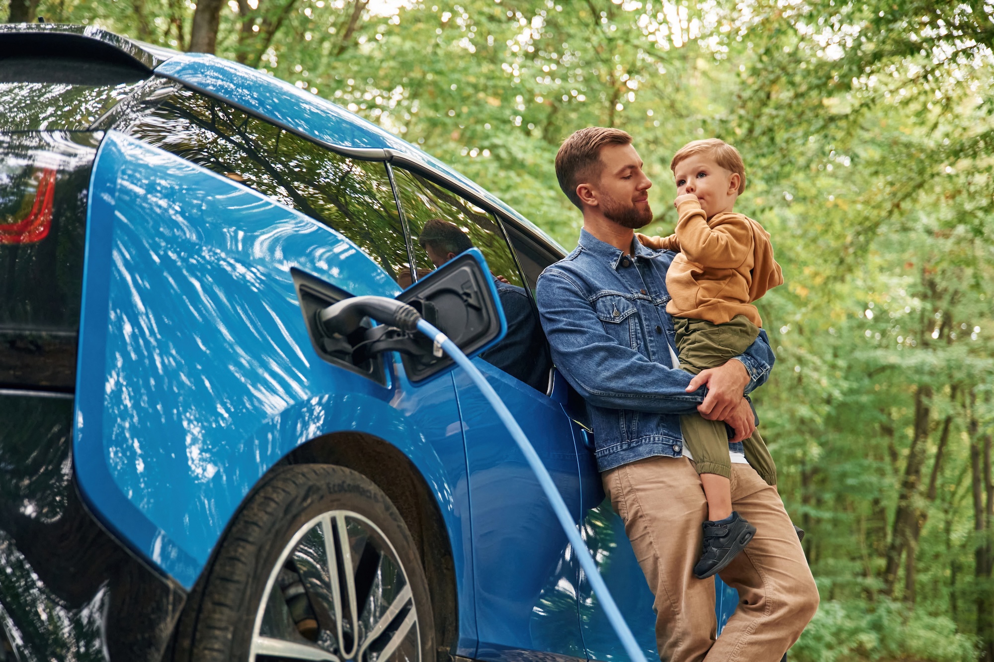 Holding Boy In Hands. Father With His Little Son Are Waiting For Electric Car To Charge